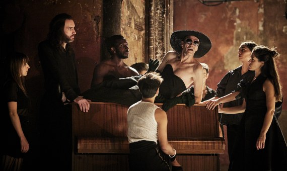 comédien allongé sur un piano et portant un chapeau noir entouré d'autres comédiens